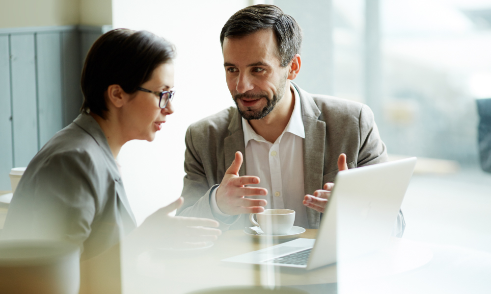 Two professionals in business attire discuss financial strategies over coffee in a modern setting, with a laptop open. This represents a family office wealth management discussion, emphasizing strategic financial planning, tax efficiency, and investment execution for high-net-worth individuals.